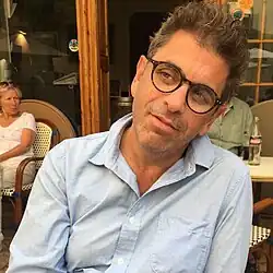 Photographie couleur montrant un homme assis à la terrasse d'un café, vêtu d'une chemise bleu au col ouvert et portant des lunettes.