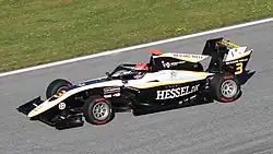 Photographie d'une monoplace de Formule 3 blanche et noire avec des traits jaunes, vue de trois-quarts gauche, d'en haut, sur une piste sèche.