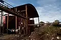 Extérieur d'une usine rouillée, le sol jonché de déchets, des habits suspendus à l'extérieur sous des réservoirs.