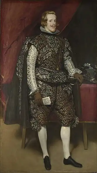 Le roi d'Espagne debout, vêtu de noir et d'argent dans une scène d'intérieur du palais.