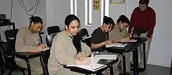 Quatre détenues en uniforme beige sont assises à des bureaux. Un professeur surveille l'une d'entre elles.