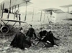 Photographie noir et blanc de quatre hommes assis dans l'herbe, en uniforme, devant un biplan.