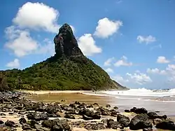 Au premier plan, une mer légèrement agitée à droite et une plage jaune recouverte de pierres noires à gauche. À l'arrière plan, une montagne recouverte d'une végétation verte de laquelle émergent des pans de roche noire.