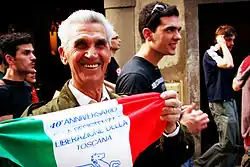 Un homme âgé souriant dans un défilé, tenant un drapeau italien décoré.