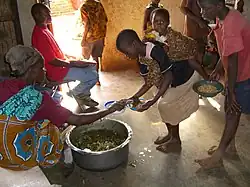 Dans une case, des enfants présentent leur assiette à une femme assise près d’un récipient métallique cabossé, posé à même le sol, et rempli d’une sorte de purée de légumes verts et orange.