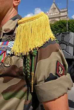 Le trèfle et la cordelette sont passés dans un bouton cousu à 1&nbsp;cm de la couture de la patte d’épaule de l’uniforme (côté gauche), la tresse passant sous l’aisselle.
