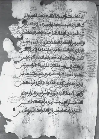 Description d'image : Photographie en noir et blanc prise par A.D.H. Bivar à la fin des années 1950, représentant une page du manuscrit connu sous le nom de 'Coran de Geidam' (manuscrit 1YM). La page contient les versets 20 à 25 de la sourate al-Baqara en écriture arabe clairement lisible, enrichie par de nombreuses annotations manuscrites en vieux-kanembu. Le parchemin est usé, avec des bords irréguliers témoignant de son ancienneté.