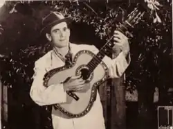 Photographie en noir et blanc d'un homme qui joue de la guitare. Il est vêtu de blanc et porte un chapeau. Derrière lui pousse des arbres.