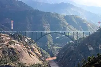 Grand pont métallique constitué d'une seule arche, franchissant une profonde vallée dans un paysage naturel de montagnes.