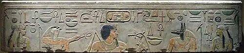 Pierre en grès clair avec des images gravées et peintes montrant deux femmes, un dieu à tête de faucon, un homme aux cheveux noirs avec une longue barbiche, un dieu à tête de chacal et des hiéroglyphes égyptiens inscrits au-dessus.