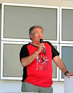 Photo en couleur d'un homme, debout, parlant devant un micro qu'il tient de la main droite.