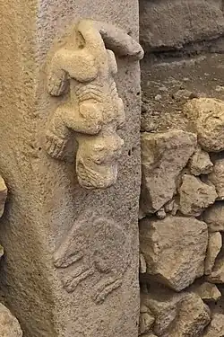 Pierre levée surmontée d'une autre. Les deux pierre forment un T et celle du dessous est partiellement enfoncée dans un mur en escalier de deux marches. Un animal est gravé en ronde bosse sur la pierre du dessous.