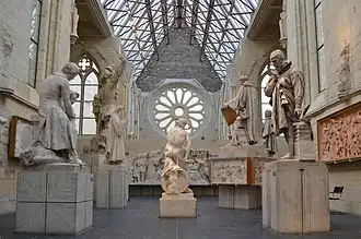  Photographie montrant plusieurs sculptures et statues dans une ancienne église ; toiture en verre et rosace.