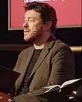 Homme brun parlant dans un micro serre tête, portant une veste de costume marron.