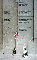 Photo agrandie d'un pan de mur gris avec des inscriptions noires, composé de plaques plus ou moins enfoncées. Des peluches et des fleurs se trouvent à son pied.