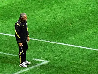 Sur une pelouse de football, un homme debout habillé en noir, est vu de profil.