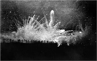 Photo noir et blanc d'un tir de nuit, gerbe de feu éclairant des barbelés.