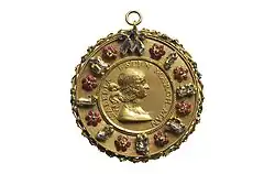 Photograhie d'une médaille dorée portant divers pierreries sur son pourtour et un portrait gravé de femme en son centre.