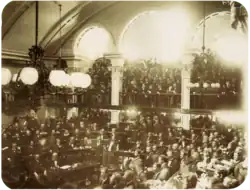 Une photo montrant un grand nombre d'hommes assis sur des gradins en demi-cercle dans une salle voûtée alors qu'une grande foule regarde d'un balcon à arcades
