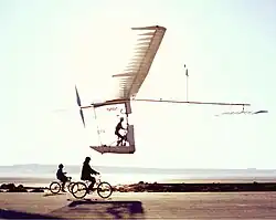 Photo de l'Albatross en vol : l'engin est conçu avec des tubes et les surfaces sont entoilées. On distingue la silhouette du pilote qui pédale. Il est à environ deux mètres du sol, suivi par deux personnes à vélo.
