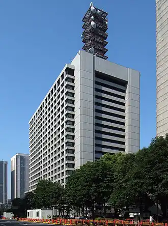 Photo d'un immeuble de bureaux d'une vingtaine d'étages, vu de 3/4 droit. La rue au pied de l'immeuble est bordée par une rangée d'arbres. L'mmeuble est surmonté d'une tour d'antennes de sept étages