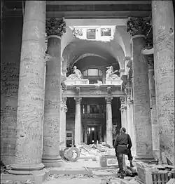 Photo noir et blanc, prise le 3 juillet 1945 à Berlin, montrant l'intérieur d'un hall du Reichstag en ruines. Au premier plan, un soldat, debout et de dos, regarde les innombrables graffitis écrits sur les colonnes de couleur claire du lieu.