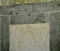 Photographie d'un panneau en marbre blanc, bordé de bandeaux verts, qui se trouve à gauche de la niche du mihrab. Il est sculpté d'un arc recti-curviligne, dont l'un des écoinçons est orné d'une inscription coufique rayonnante. Celle-ci est inscrite dans un cercle orné.