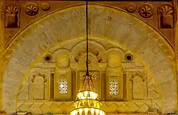 Photographie de la décoration du tympan de l'arc qui se trouve au-dessus du mihrab. Le tympan est occupé par cinq niches de différentes tailles, dont celle du milieu est la plus grande. La décoration de ce dernier inclut des carrés posés sur la pointe, des arcs à cinq lobes et des claustras garnis de verres de couleur.