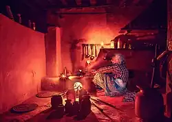 Dans une photo aux tons chauds, une dame âgée surveille, assise, la cuisson dans un four en terre.