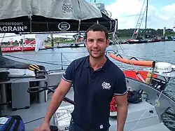 Le navigateur en buste, polo bleu marine, à l'arrière de son bateau au ponton.