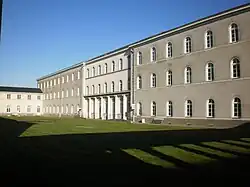 Long bâtiment blanc et rectangulaire, percé de nombreuses fenêtres, et haut de trois étages. La cour intérieure est gazonnée, on voit l'ombre d'une galerie avec colonnade, l'ensemble se dégage sur fond de ciel bleu.
