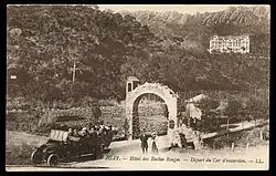 Carte postale noir et blanc. Une voiture à plusieurs places franchit un porche. Dans le lointain une batisse.