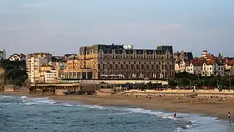 Vue d'un grand immeuble à trois étages ; façades couleur brique ou pierre et brique ; toiture en ardoises.