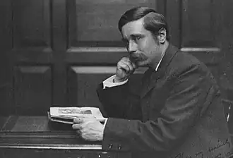 À un bureau, homme de profil moustachu, avec une raie, assis. Main droite soutient visage, gauche tient manuscrit.