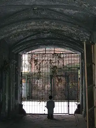 Photo en couleurs prise dans le hall d'un des bâtiments de l'ancien ghetto de Varsovie en 2013. Sous un porche de pierre peu éclairé, on distingue la silhouette d'un homme agenouillé, de dos, devant une large et haute porte grillagée donnant sur une ruelle au sol très clair. Il s'agit d'une statue en cire, de l'artiste italien Maurizio Cattelan, représentant Adolf Hitler en costume gris, priant agenouillé.