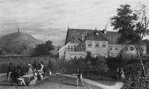 Image en noir et blanc, vaste maison en arrière-fond droit, un mont en arrière-fond gauche, au premier plan une dizaine d'hommes et de femmes mangent dans le jardin