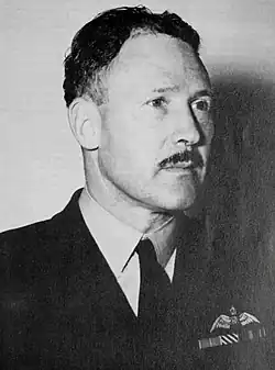 Portrait d'un homme moustachu en uniforme militaire sombre avec des ailes de pilote sur la poche gauche.