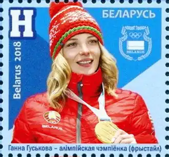 Timbre avec un fond bleu représentant une femme blonde vêtue d'un bonnet et d'un manteau principalement rouge et tenant une médaille d'or accroché autour de son cou.