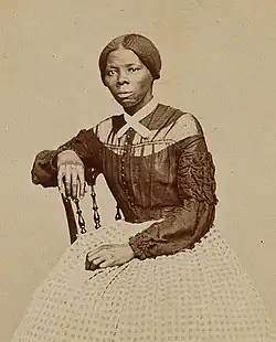 Photographie noir et blanc d'une femme noire élégante assise, le bras posé sur le dossier d'une chaise. Elle est coiffée de deux bandeaux