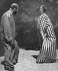 Photo noir et blanc d'un homme de dos en longue robe unie et d'une femme de face, souriante en longue robe rayée
