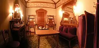 Petit salon dans lequel se trouve une table au centre, une chaise et une cheminée sur la gauche, une porte entourée de deux meubles au fond et, sur la droite, un sofa surmonté d'un miroir ainsi qu'un paravent, le tout dans le style art nouveau