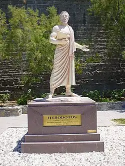 Statut blanche d'un homme barbu, portant une toge et un livre, debout sur un piédestal gris, portant l'inscription "Herodotos" sur une plaque dorée.