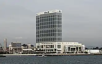 Photographie en couleurs représentant un bâtiment blanc aux nombreux étages, à la façade vitrée, sur le front de mer.