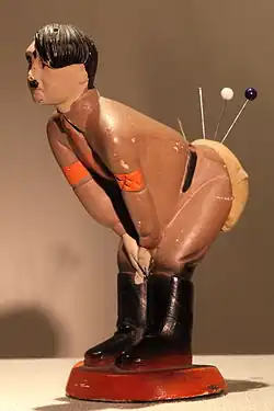 Une photo en couleurs (fond brun clair) d'une statuette en plâtre d'Adolf Hitler, posée sur un support beige. L'homme, de profil, tête nue, porte un uniforme marron clair et des bottes noires. Il est penché vers l'avant (de droite à gauche), les deux mains réunies entre ses genoux. Son derrière est recouvert d'une capote en laine orange dans laquelle sont plantées quatre épingles.