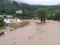 Rivière en crue, au creux d'une vallée et bordée de maisons inondées ; en arrière-plan, une colline couverte de forêt.