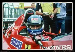 Photographie d'un pilote casqué, vu de face, dans sa monoplace de Formule 1 rouge, en gros plan.