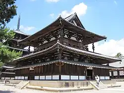 Photo couleur montrant, dressé sur une dalle en pierre beige, un bâtiment en bois à deux étages d'un temple bouddhique (couleurs dominantes de la structure : marron foncé et blanc). À l'arrière-plan, à gauche, une pagode à cinq étages, en bois marron, sur fond de ciel bleu parsemé de quelques nuages.