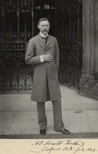 photographie noir et blanc : portrait en pied d'homme moustachu devant une grille de fer forgé
