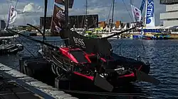 Vue de trois quarts arrière d'un voilier noir et rose, qui a la particularité d'avoir un cockpit entièrement recouvert et fermé.