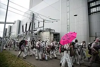 Des gens habillés en blanc et masqués pour ne pas être reconnus manifestent devant un bâtiment gris relié à des lignes électriques, probablement la centrale thermique.